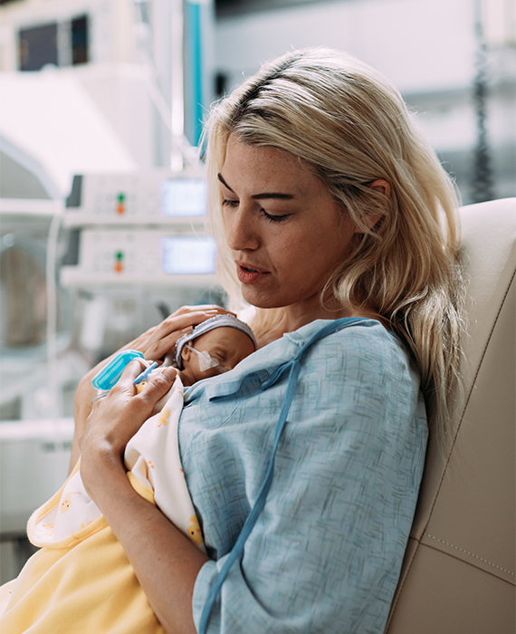 A mother holding her premature infant to her chest and looking fondly down at the baby, with Infusion pumps in the background.